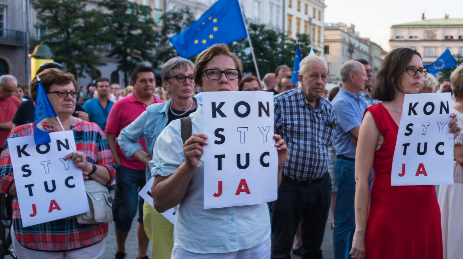Protesters are holding signs that read “KONSTYTUCJA,” which is the Polish word for “Constitution.” Some of the signs have certain letters highlighted, which is a common symbol used in Polish protests advocating for the defense of the constitution.